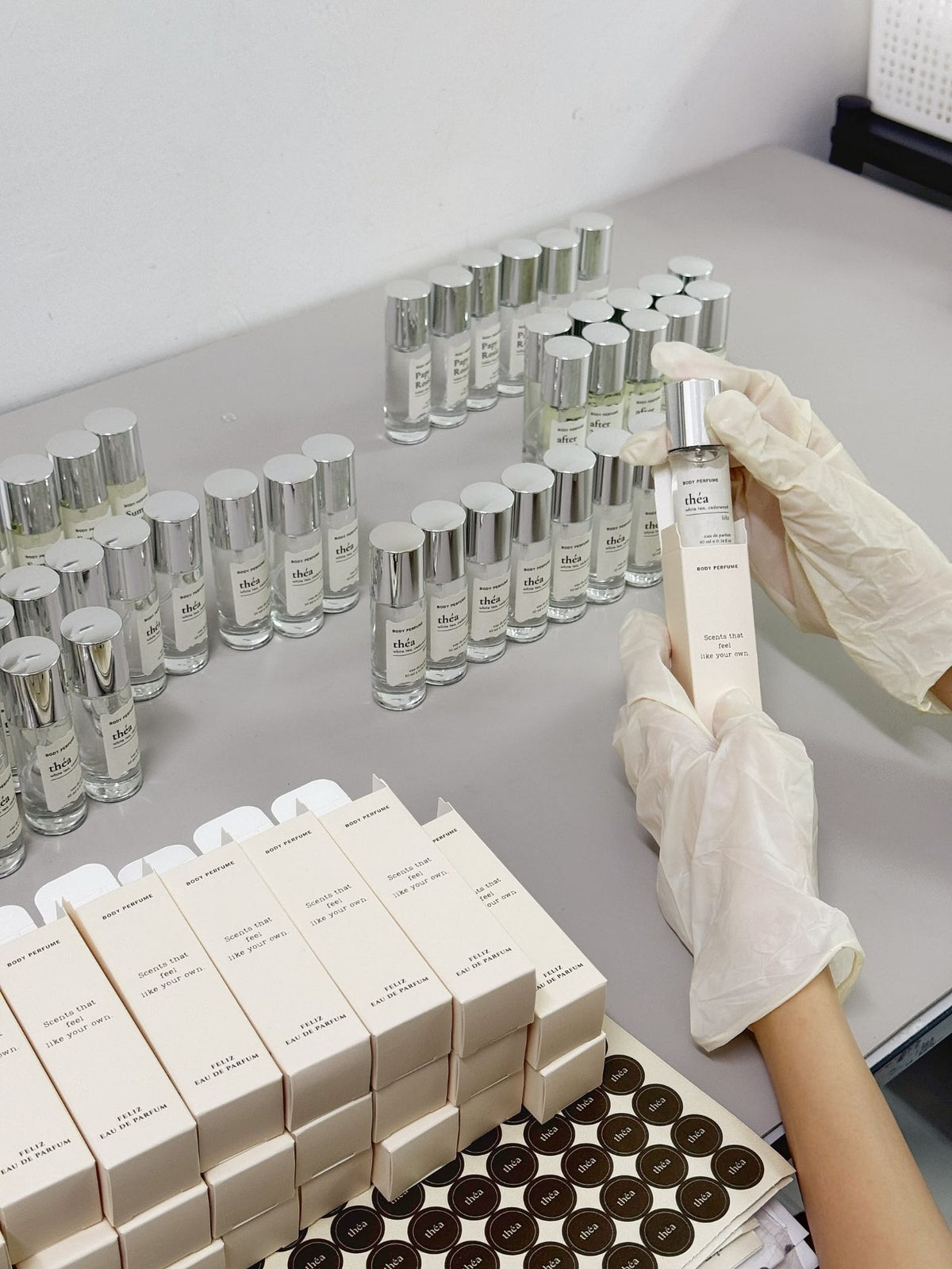 Gloved hands boxing a 10ml théa body perfume, with rows of Feliz bottles, boxes and théa stickers on the table
